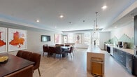 This contemporary single-floor amenity space is thoughtfully designed to accommodate both casual gatherings and structured events. The open-plan layout is anchored by large-format porcelain tile flooring that flows seamlessly throughout, lending a cohesive and polished aesthetic.

The fully equipped kitchen is a standout feature, boasting dark espresso cabinetry with brushed gold hardware, a striking geometric green hexagonal tile backsplash, stainless steel appliances including a built-in refrigerator and wall oven, and an expansive white quartz island topped by elegant globe pendant lights. The kitchen transitions naturally into the main lounge area, where a lengthy white quartz countertop bar provides additional workspace and casual seating.

The dining zone accommodates a large dark wood table surrounded by rich brown leather chairs, positioned beneath recessed lighting and adjacent to a wall-mounted flat-screen television. Textured grasscloth-style wall panels add warmth and dimension to the space. Curated contemporary artwork punctuates the walls throughout.

A dedicated game area features a foosball table and additional seating, bathed in natural light from multiple windows with horizontal blinds. A glass partition wall elegantly separates zones while maintaining visual openness. Lush tropical plants and bold abstract art complete the refined yet inviting atmosphere of this well-appointed communal space.