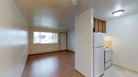 This single-floor unit offers a straightforward, functional layout with a clean and neutral aesthetic throughout. The living area is open and bright, benefiting from a large window that lets in natural light and provides views of the surrounding greenery. Warm laminate wood flooring extends through the main living spaces, adding a cohesive and inviting feel, while a baseboard heating system runs along the exterior wall for efficient climate control.

The kitchen is designed in a galley configuration, featuring warm-toned wood cabinetry — both upper and lower — paired with light laminate countertops. White appliances, including a refrigerator, electric range, and a stainless steel sink, are neatly arranged along both sides, maximizing functionality within the space. A pass-through opening connects the kitchen to the living area, promoting an open, communicative flow between the two spaces.

The bedroom is a quiet, well-proportioned room with the same laminate flooring and ample wall space. The full bathroom includes a single-sink vanity with wood cabinetry, a toilet, and a bathtub/shower combination, finished with light tile flooring for a clean, practical look. Overall, the unit presents a simple, well-maintained living environment with consistent finishes and a practical floor plan.