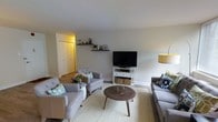 This single-floor space centers around a thoughtfully designed living room that balances comfort and style. The room is finished with light grey walls and white trim, creating a clean, neutral backdrop that enhances the sense of openness. Smooth white ceilings contribute to the airy feel of the space.

The living area is furnished with a coordinated seating arrangement, including a sofa and two tufted accent chairs in a soft grey upholstery, accented with teal and patterned throw pillows that add a touch of color. A round wooden coffee table sits atop a woven natural-fiber area rug, grounding the seating group with warmth and texture. A white media console with wicker basket storage houses a flat-screen television, while floating wooden shelves above display small decorative items and greenery.

A floor lamp with a warm yellow shade provides ambient lighting, complemented by natural light filtering into the space. Hardwood flooring extends throughout, visible beyond the rug and into an adjacent hallway, where built-in white closet doors are visible. The overall ambiance is casual yet polished, offering a practical and inviting everyday living environment.