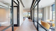 Located at 2141 Waterside Drive in Nashville, TN, this thoughtfully designed single-floor space offers a generously sized, contemporary environment ideal for gathering, collaboration, and everyday amenities. The architectural design is defined by bold black-framed floor-to-ceiling glass walls and partitions that create a sense of openness while elegantly delineating functional zones throughout.

The main gathering and dining area is a standout feature, anchored by a substantial live-edge wood table surrounded by modern seating and illuminated by a dramatic pair of sculptural branch-style chandeliers. Expansive windows flood the space with natural light and frame views of the surrounding landscape, while a built-in window-seat lounge nook provides a relaxed retreat with a patterned area rug adding warmth underfoot.

Glass-enclosed conference and meeting rooms line the corridor, offering transparency and accessibility while maintaining acoustic separation. A dedicated workstation nook features a wall-length marble-surface desk, writeable wall panels, and patterned accent wallcovering — ideal for individual focus work.

The kitchen is outfitted with sleek navy cabinetry, marble countertops, stainless steel under-counter appliances, an integrated display screen, and a textured metallic tile backsplash. A wide, tile-floored hallway connects all areas, reinforcing the clean, modern aesthetic throughout this impressive space.