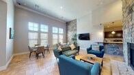 This single-floor communal space is designed to impress, combining sophisticated architectural details with comfortable, functional furnishings. The centerpiece of the main lounge is a dramatic floor-to-ceiling natural stone fireplace that anchors the room and draws the eye upward to the soaring ceilings. Large multi-pane windows line the walls, flooding the interior with generous natural light and offering views of the surrounding outdoors. The lounge area features a curated seating arrangement of teal velvet sofas and bold blue accent chairs, complemented by a mid-century modern coffee table and warm tile flooring throughout. A dedicated dining nook with round marble-top tables and upholstered chairs provides a comfortable space for casual gathering or meetings. A wall-mounted flat-screen television is positioned opposite the fireplace for easy viewing. Adjacent to the main lounge, a well-appointed kitchenette offers dark wood cabinetry, granite countertops, a stone-accented breakfast bar, appliances including a microwave and coffee maker, and pendant lighting overhead. A hallway connects additional rooms, which appear to include a game room with recreational equipment visible in the background. The overall ambiance is warm, welcoming, and stylishly appointed.