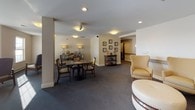 This single-floor communal lounge is thoughtfully designed to accommodate a variety of social activities and relaxed gatherings. The space is organized into distinct seating zones that flow naturally into one another, unified by consistent neutral-toned carpeting and clean white and beige walls. Recessed lighting throughout provides even, comfortable illumination, complemented by warm accent lamps in the activity areas.

A central lounge area features a collection of upholstered wingback chairs in cream and patterned gold fabric, arranged around a tufted round ottoman, creating an inviting conversational setting. Structural columns subtly define zones within the open floor plan without interrupting the sense of spaciousness.

A secondary area accommodates a game or activity table surrounded by wood-framed chairs, accompanied by a sideboard with decorative lamps and accessories. Framed artwork along the walls adds visual interest, while a console table with decorative accents contributes to the space's refined character. Large windows allow natural light to filter in, offering views of the surrounding building exterior. Multiple doorways connect to adjacent corridors, ensuring easy accessibility and circulation throughout the space.