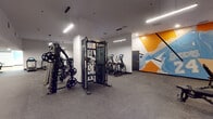 Located on Wilshire Boulevard in the heart of Los Angeles, this single-floor fitness center offers a well-appointed and modern workout environment designed to support a full range of exercise activities. The main gym area features durable rubber flooring and is equipped with a functional training cable machine loaded with weight plates, along with an array of cardio equipment including treadmills and elliptical machines with digital displays. Sleek linear pendant lighting illuminates the space from above, complementing the clean white walls. A vibrant abstract mural in orange, blue, and white adds a dynamic, energizing element to the gym floor, giving the space a contemporary and motivating atmosphere. An additional area visible through an open passage offers supplemental workout space with light wood flooring, providing flexibility for varied training needs. A dedicated studio room rounds out the facility, featuring a smooth hardwood floor ideal for group fitness classes, yoga, or dance. This studio is lined with full-length mirrors along one wall, fitted with acoustic panel tiles for sound management, and decorated with a bold geometric mural in teal, yellow, and light blue. Recessed and flush ceiling lights keep the studio bright and functional. Together, these spaces create a comprehensive, thoughtfully designed fitness facility.