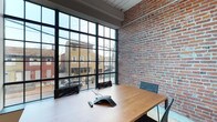 Roebling Lofts is a thoughtfully designed single-floor space that masterfully combines raw industrial heritage with contemporary functionality. The space is defined by its striking exposed brick walls, which lend warmth and texture throughout, complementing the clean lines of the modern interior. Floor-to-ceiling steel-framed grid windows flood the space with abundant natural light while offering open views of the surrounding urban streetscape, creating a seamless connection between the interior and the neighborhood outside. The conference area features a generous wood-topped meeting table surrounded by sleek task chairs, equipped with integrated conferencing technology — making it well-suited for professional gatherings and collaborative work sessions. The interplay between the rugged brick, the dark steel window frames, and the warm wood tones creates a cohesive and inviting atmosphere that balances character with practicality. Exposed structural elements overhead further reinforce the loft's authentic industrial origins. Roebling Lofts offers a distinctive and inspiring environment that honors its architectural roots while meeting the demands of a modern workspace.