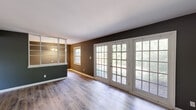 Located at 1909 Hazelwood Ave in Fort Wayne, Indiana, this one-bedroom, one-bathroom home offers a well-organized single-floor layout with thoughtful design details throughout. The combined living and dining area is a standout feature, anchored by a striking wall of white-framed French doors that flood the space with natural light and provide views of the surrounding greenery. Light-toned hardwood-style flooring runs through the main living areas, complementing the neutral gray and taupe wall tones that lend a cohesive, modern feel. The dining area is defined by an elegant chandelier and large windows, with built-in open shelving adding both function and character. The kitchen is efficiently arranged with white cabinetry, light countertops, and black appliances including a range and refrigerator, offering ample storage for its scale. The bedroom features plush carpeting, a natural window for daylight, and a sliding-door closet providing practical storage. The bathroom is well-appointed with full-wall warm-toned tile surrounding a tub-shower combination, a marble-topped vanity, and a window for natural ventilation. A connecting hallway ties each room together, creating a logical and easy-to-navigate flow throughout this compact yet complete home.
