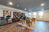 Located at 7200 Chase Oaks Blvd in Plano, Texas, this single-floor community amenity space is thoughtfully designed to serve multiple functions in a cohesive, stylish environment. Throughout the entire space, striking herringbone-patterned hardwood flooring creates a refined visual continuity that anchors each area's distinct purpose.

The main lounge and library area features a dramatic dark ceiling with recessed lighting, built-in dark wood bookshelves stocked with books and decorative objects, and a comfortable seating arrangement of upholstered accent chairs in warm amber and neutral tones set atop a colorful area rug. A dedicated workstation with a computer and printer is tucked along one wall, while large windows along an adjacent wall invite abundant natural light and exterior views.

A connected multi-purpose meeting room provides a more structured setting, outfitted with rectangular tables and cushioned chairs arranged for group gatherings. Warm botanical artwork adorns the clean white walls, and the same dark ceiling treatment maintains design consistency throughout.

The kitchenette is a functional and polished space featuring full-length gray shaker cabinetry with gold hardware, dark countertops, a matte black faucet, a single-basin sink, and a herringbone tile backsplash that echoes the flooring motif. This well-equipped area supports catered events and everyday community use with ease.