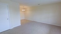 This single-story space presents a straightforward and functional layout designed to maximize usable living area. The interior is finished with neutral, light beige walls that lend a calm and understated ambiance throughout. Plush wall-to-wall carpeting in a complementary warm tone covers the floors, providing comfort underfoot and contributing to a cohesive aesthetic.

The main living area is generously proportioned, offering an open feel with clearly defined spaces. A partial wall creates a natural division between areas, subtly separating the rooms while maintaining an open connection between them. A classic multi-arm chandelier serves as a focal point in the adjoining area, adding a touch of traditional elegance to the otherwise minimalist interior.

White baseboards line the perimeter of the rooms, providing a clean contrast against the carpeted floors. Ceiling-mounted HVAC vents are integrated neatly into the design, ensuring efficient climate control. Electrical outlets are conveniently positioned along the walls. The overall palette and finishes create a neutral canvas that is highly adaptable to a variety of furnishing styles and personal preferences.