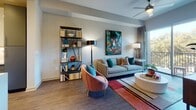 This thoughtfully designed single-floor residence offers a seamless open-concept layout that connects the kitchen, dining, and living areas into one cohesive, functional space. The kitchen is a standout feature, showcasing two-tone cabinetry with sleek white upper cabinets and muted grey lower cabinets, quartz countertops, stainless steel appliances, and a generous island with an integrated sink and bar seating — all illuminated by elegant glass pendant lights and a track lighting system overhead. Rich dark hardwood-style flooring runs throughout the main living areas, grounding the space with warmth and contrast. The living room is bright and inviting, benefiting from abundant natural light streaming through floor-to-ceiling sliding glass doors that open to a private balcony with leafy outdoor views. A tray ceiling with a ceiling fan adds architectural interest to the bedroom, which features a large window framing treetop views and creating a peaceful, light-filled retreat. The bathroom is finished with large-format marble-look wall tiles, mosaic floor tiles, and a frameless glass-enclosed walk-in shower, lending a clean and spa-like aesthetic. Colorful contemporary artwork displayed throughout adds a vibrant, curated character to the overall ambiance of this well-appointed residence.