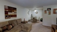 This single-floor residence offers a welcoming and functional layout designed for comfortable everyday living. The combined living and dining area serves as the heart of the home, bathed in soft, neutral tones that create a calm and inviting atmosphere throughout. The living area is anchored by a plush sectional sofa in warm beige tones, complemented by a small side table and tasteful wall art that adds a touch of personality to the space. An ornate wall clock serves as a subtle decorative focal point. The dining area sits just beyond the living space, featuring an elegant glass-top table surrounded by wicker-style chairs, lending the room a light and airy feel. A ceiling fan overhead provides both practical ventilation and gentle ambient lighting for the combined space. The flooring throughout is finished in neutral carpet, contributing to the cozy and understated character of the home. A baseboard heating unit runs along the wall, ensuring year-round comfort. Subtle greenery in the corner adds a natural element to the interior. The overall design is clean, straightforward, and functional, making this a practical and comfortable single-floor living space.