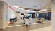 This single-floor space, branded "ZEN," presents a refined, contemporary design that blends functionality with upscale aesthetics. The entrance lobby makes an immediate impression with large-format porcelain tile flooring, a sculptural geometric pendant light fixture, and a striking feature wall combining warm wood paneling, textured stone, and the bold ZEN logo medallion. Planters with ornamental grasses add a natural, calming touch to the entryway.

Beyond the lobby, wide corridors clad in light wood paneling lead into a spacious communal lounge area. The lounge features an architecturally detailed ceiling with layered wood beam accents and integrated recessed lighting, creating depth and visual interest overhead. Wide-plank wood-look tile flooring runs throughout, anchoring the neutral, warm color palette.

The lounge is thoughtfully divided into multiple seating zones, including round marble-topped tables paired with upholstered chairs on gold metal frames, and a dedicated bar counter with dark cabinetry and navy blue bar stools. A full kitchen area with pendant lighting and built-in appliances supports catering and entertaining needs. Floor-to-ceiling glass doors along one wall allow natural light to flow in and provide views to an exterior terrace, enhancing the overall sense of openness and airiness throughout the space.