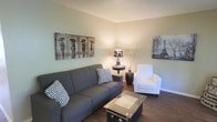 This single-floor space features a thoughtfully designed living area that balances comfort and contemporary style. The living room is anchored by a charcoal grey sofa adorned with geometric patterned throw pillows, complemented by a clean white accent chair that adds contrast to the room. Dark hardwood flooring runs throughout, grounding the space with warmth and visual depth. The walls are painted in a soft sage green, creating a calming, neutral backdrop that pairs well with the understated decor. A textured popcorn ceiling adds a classic architectural detail overhead. Decorative wall art, including a rustic multi-figure painting and a Parisian-themed canvas print, contribute personality and character to the room. A small round side table with a stylish patterned lamp and dried branch arrangement provides ambient lighting and an organic decorative touch. The vintage-style trunk coffee table serves as a functional yet distinctive centerpiece. Overall, the space presents a cohesive, relaxed ambiance with carefully curated furnishings and decor that make it feel welcoming and well-appointed.