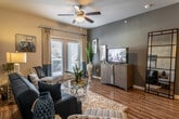 This single-floor residential unit offers a thoughtfully designed layout that balances functionality and comfort across its key living spaces. The kitchen is a standout feature, showcasing rich warm-toned wood cabinetry extending to the upper walls, granite countertops with decorative stone tile backsplash, black appliances including a refrigerator and range, and wood-look flooring. Track lighting illuminates the galley-style workspace, which opens toward a dedicated dining area with a glass-top table. The living room is bright and inviting, centered around a ceiling fan with light fixture and anchored by wood-look flooring. French doors along one wall invite natural light and offer access to the outdoors, creating an airy, open ambiance. Bold accent walls in deep charcoal tones add a contemporary edge to the entryway and main corridor, which features hardwood-style flooring and a granite-topped bar counter. The bedroom provides a calm retreat with plush carpeting, a ceiling fan, and natural light filtering through window blinds. Neutral wall tones complement the room's understated décor, which includes a dresser, bedside tables, and decorative wall art. The space as a whole presents a cohesive design with a warm, livable aesthetic throughout.
