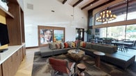 This single-floor space makes a strong first impression through its dramatic vaulted ceiling adorned with dark, exposed wooden beams that contrast beautifully against crisp white walls. The generous ceiling height amplifies the sense of openness throughout the room. A large-scale, vibrant portrait artwork anchors one wall, framed by warm wood paneling that adds a grounding, organic element to the interior. The main seating area centers around a substantial gray sectional sofa layered with an eclectic mix of earth-toned and jewel-colored throw pillows, paired with a geometric, faceted wood coffee table that serves as a sculptural focal point. A cognac leather accent chair adds a rich contrast to the neutral palette. The space is further furnished with a round side table and additional seating visible near floor-to-ceiling windows, which flood the area with natural light and offer views of lush greenery outside. An elegant chandelier with warm-toned bulbs illuminates a dining or secondary seating area in the background. The layered area rug, mix of textures, and curated décor collectively create a sophisticated yet welcoming ambiance.