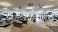 This spacious fitness center offers an impressively open and well-organized layout designed to accommodate a full range of workout needs. The interior is finished with neutral gray walls and durable patterned tile flooring, creating a clean and professional atmosphere throughout. Multiple ceiling fans are distributed across the space to maintain airflow and comfort during exercise.

Natural light streams in through numerous windows fitted with white blinds, providing views of surrounding greenery and keeping the environment feeling bright and welcoming. Large floor-to-ceiling mirrors line key sections of the walls, amplifying the sense of space and allowing users to monitor their form during training.

The cardio zone features a row of treadmills equipped with individual screens, alongside elliptical trainers and stationary bikes, including recumbent models. A dedicated strength training area houses a variety of cable machines and multi-functional cable systems, including Hoist-brand equipment, complemented by a full dumbbell rack and adjustable benches. Protective rubber flooring mats are positioned beneath the free weight stations for safety and durability.

Additional amenities include wall-mounted water fountains, a waste station, and pendant and overhead linear lighting for bright, even illumination. A counter area with seating is visible near the entrance, adding a functional lounge element to the facility. The overall ambiance is modern, clean, and purpose-built for serious fitness use.