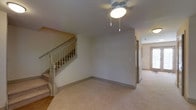 This two-floor residence offers a functional and well-structured layout designed for comfortable everyday living. The main level features a spacious carpeted living area illuminated by a ceiling fan light fixture, providing both practical lighting and air circulation. Neutral-toned walls throughout create a clean, versatile backdrop well-suited to a variety of interior styles. A classic staircase with turned wood balusters and a painted handrail connects the two floors, adding a traditional architectural touch to the interior. The entry area transitions seamlessly from hardwood-style flooring into the carpeted living space, providing a natural delineation between zones. A hallway leads further into the home, hinting at additional rooms and living spaces beyond. The overall color palette is warm and understated, with light beige carpeting and soft white walls contributing to a bright, airy ambiance. The consistent trim and baseboard detailing throughout reflect careful attention to finish work. Across both floors, the layout appears thoughtfully designed to maximize usable space while maintaining an open and approachable feel, making this a well-rounded property suitable for a range of occupants.