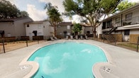 This residential community amenity space encompasses two distinct areas designed to serve and enhance daily living. The outdoor pool area features a generously sized freeform swimming pool with clear turquoise water, framed by mature oak and palm trees that provide natural shade and a lush, relaxed atmosphere. A spacious concrete deck surrounds the pool, outfitted with multiple lounge chairs and patio seating, making it ideal for leisure and recreation. An iron perimeter fence encloses the pool area, with the surrounding residential buildings adding a sense of privacy and community. The indoor mailroom is a clean, modern space finished in soft aqua-toned walls and light tile flooring that reflects natural light streaming in through large floor-to-ceiling windows. Rows of dark bronze individual mailboxes line both sides of the room, organized efficiently within contemporary dark gray cabinet structures. A centrally positioned package sorting counter with a built-in drop slot provides added convenience for package retrieval. Framed geometric artwork accents the walls, lending a polished, curated aesthetic to the functional space. Recessed and track lighting illuminate the interior evenly, creating a bright and welcoming environment for residents.