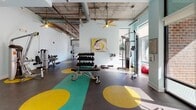 Located at 540 Selma St in Savannah, Georgia, this single-floor fitness center offers a well-equipped workout environment designed with both functionality and style in mind. The space features an industrial-chic aesthetic, highlighted by exposed concrete ceilings with visible ductwork, ceiling fans, and light blue walls that create a fresh, energizing atmosphere. Vibrant teal and yellow circular accents on the dark rubber flooring add a playful, modern touch throughout the facility.

The cardio zone is lined with a row of Cybex treadmills and elliptical machines positioned along large windows, allowing natural light to flood the space and offering views of the surrounding streetscape. Wall-mounted televisions accompany the cardio equipment for an engaging workout experience. An adjacent area houses a comprehensive free weight section, including a full dumbbell rack, adjustable benches, and cable machines from Hoist, catering to a wide range of strength training needs.

A dedicated strength zone features a full barbell squat rack with weight plates, situated alongside floor-to-ceiling glass walls that bring in ample daylight. The facility also includes additional cable and functional training machines. Natural light is abundant throughout, with multiple large windows and glass doors reinforcing an open, airy feel. The overall layout is spacious and logically organized, making it a well-rounded fitness destination.