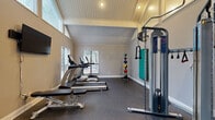 Located at 100 Ashley Creek Ct in Stone Mountain, GA, this dedicated fitness center offers a well-equipped workout space within a charming architectural shell. The room's most distinctive feature is its soaring vaulted ceiling finished with white-painted beadboard paneling, which lends a clean, airy aesthetic and creates a sense of spaciousness well beyond its compact footprint. Recessed lighting supplements the generous natural light that streams in through multiple windows fitted with horizontal blinds, providing views of the surrounding greenery.

The durable rubber flooring throughout the space is both practical and slip-resistant, ideal for active use. Equipment is thoughtfully arranged to maximize the available floor area and includes True-brand treadmills, an elliptical trainer, a multi-function cable resistance machine, a dumbbell rack with a full set of free weights, a weight bench, and a collection of medicine balls mounted on a wall rack. A wall-mounted flat-screen television adds an entertainment option for cardio sessions. Entry is through a stylish black-framed glass door that connects to an adjacent corridor. Neutral warm-gray walls complete the clean, functional aesthetic of this well-appointed fitness facility.