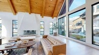 Located at 212 E 1720 N in Heber City, Utah, this single-floor space delivers an impressive blend of modern alpine architecture and warm, natural materials. The open living area is immediately defined by its most striking feature: a soaring vaulted ceiling clad in rich, warm-toned wood planking and supported by exposed structural beams, creating a dramatic sense of verticality and openness. Crisp white walls provide a clean contrast to the natural wood overhead, while light hardwood flooring runs throughout, reinforcing the airy, cohesive aesthetic. A bold, white statement fireplace anchors the main living area, serving as a sculptural focal point with a built-in log storage niche beneath. Expansive floor-to-ceiling windows and oversized black-framed glass panels line the walls, flooding the interior with abundant natural light and offering unobstructed views of the surrounding Heber Valley neighborhood and the mountain ridgeline beyond. A separate nook area features large picture windows, a marble-topped table, and a built-in bench, making it an ideal spot for casual seating. Custom wood cabinetry is integrated seamlessly into the architectural design. The overall ambiance is bright, inviting, and refined, balancing mountain warmth with contemporary minimalism.