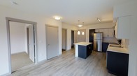 This single-floor, one-bedroom, one-bathroom unit offers a thoughtfully designed open-concept layout that maximizes functionality. The kitchen serves as a focal point, featuring dark cabinetry contrasted by a light countertop island, stainless steel refrigerator, and upper white cabinets. Pendant lighting above the island adds a modern touch, complementing the track lighting nearby. Light-toned laminate wood flooring runs throughout, lending warmth and visual continuity to the space. The open layout connects the kitchen seamlessly to the living area, creating an efficient and inviting flow. Multiple doorways visible from the main living space lead to the bedroom, bathroom, and closet areas, all framed by clean white walls and contemporary door hardware. Flush-mount ceiling lighting provides ample illumination throughout. The overall aesthetic is clean and modern, with a neutral color palette of white, grey, and dark navy tones that creates a balanced, contemporary ambiance well-suited for comfortable everyday living.