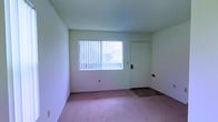 This single-story space presents a straightforward, functional layout with clean, white-painted walls throughout. The room visible in the image is finished with wall-to-wall carpet in a muted rose-pink tone, providing a soft underfoot feel. A large window dressed with vertical blinds serves as the room's primary source of natural light, allowing diffused brightness to fill the interior. The textured ceiling adds a modest layer of detail overhead, while a fire sprinkler head is centrally positioned. A solid entry door equipped with a keypad lock offers convenient, secure access. The transition area near the door features a hard flooring surface, contrasting with the surrounding carpet and providing a practical entryway zone. The overall ambiance is understated and neutral, offering a blank canvas that lends itself well to a variety of uses or furnishing styles.