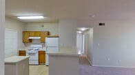 This single-floor residential unit offers a functional and well-organized layout with a clear separation between living, sleeping, and kitchen areas. The kitchen is equipped with warm honey-toned wood cabinetry, white appliances including a refrigerator, electric range, and dishwasher, as well as a double-basin sink and light-colored countertops. A peninsula counter provides additional workspace and serves as a natural divider between the kitchen and the adjoining living area. The kitchen features tile flooring and is illuminated by overhead fluorescent lighting, with ventilation provided through ceiling vents. The living area is carpeted and receives natural light through a window and a glass-paneled exterior door, giving the space an open, airy feel. A ceiling fan with a light fixture keeps the area comfortable. The bedrooms are also carpeted and feature ceiling fans, neutral-toned walls, and multiple windows fitted with blinds that allow for adjustable natural light. The bathroom includes a full tub and shower combo with a classic white tile surround accented by a decorative taupe border tile, complemented by chrome fixtures. Overall, the space presents a clean, neutral palette with practical finishes throughout.