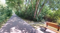 Rails To Trails is a well-established recreational corridor that repurposes former railway infrastructure into a multi-use paved path for walkers, joggers, and cyclists. The trail stretches through a lush, tree-lined corridor where mature deciduous trees form a natural canopy overhead, casting dappled shade across the smooth asphalt surface. Dense shrubs and leafy undergrowth flank both sides of the path, creating a sense of immersion in nature while still maintaining a clear, accessible route for users of all abilities. Wooden benches are positioned along the trail, providing convenient rest stops and opportunities to take in the surrounding greenery. In areas where the canopy opens up, views extend to neighboring residential properties and open sky, offering a pleasant contrast to the shaded stretches. Supporting the trail's upkeep is a dedicated utility and storage facility, which houses plumbing infrastructure, maintenance supplies, and janitorial equipment organized on shelving units. This operational space ensures the trail remains clean and well-maintained year-round. Overall, Rails To Trails offers a tranquil, nature-rich outdoor experience that seamlessly blends accessibility with a verdant, park-like atmosphere, making it an inviting destination for community recreation and active transportation.