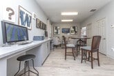 The Cyber Cafe is a thoughtfully designed single-floor space that seamlessly blends functionality with a relaxed, welcoming atmosphere. The interior features light-toned walls that contribute to a bright and open feel, complemented by natural light filtering through a window dressed with white blinds and neutral curtains.

A built-in countertop workstation runs along one wall, offering ample surface space and equipped with a desktop computer and printer, making it ideal for residents or visitors needing to handle digital tasks. Below the counter, clean white cabinetry provides generous storage while maintaining a streamlined aesthetic.

The walls are adorned with a curated selection of framed artwork, including bold typographic pieces and abstract works in dark, moody tones that add visual interest and personality to the space. A distinctive "@" symbol display nods to the digital nature of the cafe's purpose.

Adjacent to the workstation area, a comfortable seating arrangement with upholstered chairs and a round table creates an inviting lounge space, perfect for casual meetings or relaxed browsing. Overhead, flush and industrial-style lighting fixtures ensure the space remains well-lit throughout.

Overall, the Cyber Cafe strikes a balance between a productive work environment and a comfortable gathering space, making it a versatile and functional amenity.