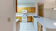 This single-story residence offers a well-organized layout with distinct living, kitchen, bedroom, and bathroom spaces. The open-concept living area features wood-look flooring and crown molding, creating a clean, cohesive aesthetic. A doorway connects the living space to the rest of the home, allowing for an easy flow throughout.

The kitchen is equipped with warm-toned wood cabinetry extending to the upper walls, light speckled countertops, and a full suite of white appliances, including an electric range, built-in microwave, and refrigerator. A peninsula with a sink provides additional counter space and casual dining potential. Light tile flooring completes the kitchen's functional design.

The bedrooms are carpeted and feature ceiling fans, neutral walls, and windows with vertical blinds that allow natural light and outdoor views. The rooms vary in size, offering flexible use options.

The bathrooms include light wood vanity cabinetry, speckled countertops, and bright vanity lighting above large mirrors. One bathroom features a tub-shower combination with white tile surround and tile flooring, while another offers a double-sink vanity with a tall storage cabinet. Overall, the residence presents a practical, well-maintained interior.