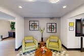 Located at 811 Harvey Rd in College Station, Texas, this single-floor common area delivers a bold and energetic atmosphere through its distinctive retro-modern design. Vivid green and olive accent walls contrast sharply with crisp white ceilings and light wood-look tile flooring, creating a lively yet cohesive visual identity throughout the space.

The room is divided into several functional zones. A well-appointed kitchenette area features dark cabinetry with a granite-style countertop, open shelving displaying decorative dishware and greenery, a beverage cooler, and a coffee station — all set against a rich green feature wall adorned with framed artwork.

An adjoining lounge area offers comfortable chartreuse armchairs arranged around a cylindrical wood coffee table atop a checkered area rug, lending a mid-century modern flair. Geometric wall-mounted décor frames the seating nook, adding visual depth.

A dedicated business center nook provides wall-mounted desks with computers and sleek green chairs alongside a flat-screen TV and stylish storage cabinet. Frosted glass French doors delineate areas while maintaining an open, airy feel. Recessed lighting throughout ensures even illumination, enhancing the space's welcoming and functional ambiance.