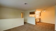 This single-floor space presents an open-concept layout that connects the living area and kitchen in a practical, flowing arrangement. The living area is finished with wall-to-wall carpeting in a neutral tone, creating a warm and understated foundation. A half-wall partition separates the living space from the kitchen, maintaining an open sightline while defining each area. The kitchen features wood-toned upper and lower cabinetry, a white electric range, and track lighting overhead, providing ample illumination for cooking and meal preparation. Additional overhead cabinetry adds storage capacity above the partition. A hallway visible from the main living area leads to further rooms within the unit, hinting at additional private spaces beyond the shared living zone. The ceiling is standard height with a central HVAC vent, contributing to a comfortable interior climate. The overall ambiance is functional and neutral, with a clean palette of white walls and beige carpeting that offers a versatile backdrop suited to a variety of furnishing styles.