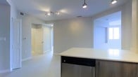 This single-floor residence offers a clean, modern aesthetic defined by an open-concept design and a neutral color palette that creates a bright, welcoming atmosphere throughout. The kitchen serves as a central highlight, featuring a large island with a crisp white countertop and a stainless steel dishwasher integrated into warm wood-toned cabinetry. Track lighting and pendant fixtures illuminate the space effectively, complementing the large-format white tile flooring that extends seamlessly across the living areas. The open layout connects the kitchen to the main living area, allowing for natural sightlines and a sense of spaciousness. A hallway leads to additional rooms, with white panel doors providing clean transitions between spaces. The walls are finished in a soft, light gray tone that reflects light well and enhances the overall airy quality of the interior. Recessed ventilation and thoughtful ceiling fixtures contribute to the polished, move-in-ready feel of the space. The combination of contemporary finishes, an efficient floor plan, and cohesive design elements throughout makes this a well-appointed and functional single-level living space.