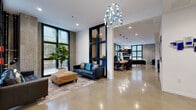 Located in Berkeley, CA, this lobby and co-working space seamlessly blends modern industrial design with comfortable, functional environments. Polished concrete floors extend throughout, reflecting light and lending the space an open, airy feel. Floor-to-ceiling black-framed windows flood the interior with natural light while offering views of lush greenery outside.

The entrance lobby features a striking lounge area with tufted leather sofas, a woven ottoman, and a lush fiddle-leaf fig tree, creating a welcoming first impression. A dramatic chandelier composed of cascading blue glass pendants serves as a standout focal point overhead.

The co-working area is anchored by a substantial reclaimed wood communal table surrounded by ergonomic chairs, lit by oversized industrial pendant lamps. Modular privacy workstations with blue divider panels provide dedicated focus areas alongside an ornate textured accent wall. Curated artwork, including colorful mixed-media pieces, is displayed throughout, adding a creative energy to the space.

A lounge nook at the far end features a fireplace, wall-mounted television, floor-to-ceiling windows, and a mix of leather and upholstered seating — ideal for informal meetings or relaxation. An elevator and accessible entry door with decorative wall sculptures complete the thoughtfully designed space, making it both professional and inviting.