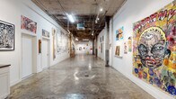 Located at 2450 Roosevelt Ave in San Antonio, TX, this single-story gallery space offers an industrial-chic environment purpose-built for contemporary art exhibition. The layout follows an L-shaped floor plan, providing two distinct gallery wings that connect through a central junction, creating a natural flow for visitors exploring the exhibitions.

The space is defined by its raw, industrial aesthetic. Polished and weathered concrete floors run throughout, complemented by exposed ceilings featuring aged structural elements, visible ductwork, track lighting, and original concrete columns. The contrast between the pristine white display walls and the unfinished ceiling gives the gallery a compelling, edgy character well-suited to its bold artwork.

The main gallery wing is generously proportioned, offering extensive wall space capable of accommodating large-scale paintings, mixed-media installations, and oversized textile works. Vibrant street-art-inspired murals and figurative works fill the walls, creating an immersive visual experience. Track and pendant lighting highlight individual pieces throughout.

A smaller connecting area near the entrance features glass-paneled partitions and a casual seating arrangement, providing a welcoming reception space. Built-in cabinetry with counter space along one wall offers practical utility for gallery operations. Additional rooms accessible via white-painted doors along the corridor provide supplementary spaces for storage or private use.