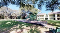This single-story residential community is set within a beautifully landscaped outdoor environment that defines its overall character and appeal. The grounds are expansive and well-maintained, featuring lush green lawns shaded by a striking collection of mature trees, including a large, sprawling banyan tree that serves as a natural focal point of the common area. Beneath its broad canopy, green park benches are thoughtfully placed, offering residents a peaceful spot to relax and enjoy the outdoors. The community's exterior buildings are finished in light, cheerful tones of green and white, consistent with a classic Florida architectural aesthetic. Balconies and walkways are visible along the multi-unit structures, adding to the open, airy feel of the property. A dedicated playground area features a robust, multi-component play structure with slides, climbing elements, and activity panels set over a protective rubberized surface — making it an ideal amenity for younger residents. The overall ambiance of the community is calm and inviting, with the natural tree canopy providing ample shade and a sense of privacy. The combination of thoughtfully designed communal spaces and lush tropical landscaping creates a comfortable and welcoming outdoor living environment throughout the property.