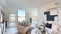 This contemporary single-floor residence showcases a thoughtfully designed open layout bathed in natural light. The living and dining area serves as the heart of the home, highlighted by large floor-to-ceiling windows that frame sweeping urban views and flood the space with sunlight. Light hardwood flooring runs throughout, complementing the clean white walls and modern aesthetic. The living area offers generous seating space, a wall-mounted flat-screen TV, and a ductless mini-split climate control unit, ensuring year-round comfort. A dedicated dining nook sits beside the expansive windows, creating an ideal setting for meals with a view. The primary bedroom is a serene retreat featuring a wall-mounted TV, a ductless climate control unit, and soft natural light filtering through a large window with frosted privacy panels. A second bedroom mirrors the minimalist design, with clean lines and ample natural light. The bathroom is finished in crisp white subway tile, featuring a glass-enclosed shower stall and light wood-tone flooring that ties seamlessly to the rest of the unit. Throughout the residence, the consistent use of neutral tones, recessed lighting, and modern fixtures creates a cohesive, sophisticated atmosphere well-suited for contemporary urban living.