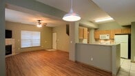 This two-floor space features an open-concept main level that seamlessly connects the living area and kitchen. The flooring throughout the main level consists of warm hardwood-style planks, lending a cohesive and inviting feel to the space. The living area is equipped with a ceiling fan with a light fixture, adding both functionality and a casual aesthetic. A large window allows natural light to fill the room, brightening the sage-green painted walls.

The kitchen is positioned adjacent to the living area and is separated by a raised breakfast bar, which provides both a casual dining option and a natural division between spaces. The kitchen features light wood cabinetry, a tile backsplash, and includes a built-in microwave and range. A pendant light fixture hangs over the breakfast bar area, adding a focal point to the space.

A door visible at the far end of the main level provides access to additional areas of the home, while a staircase leads to the second floor. The layout of the main level prioritizes open, functional living with clear sightlines across the space.