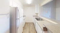 This single-floor space offers a clean and functional layout designed for comfortable everyday living. The galley-style kitchen is a standout feature, characterized by its efficient use of space with cabinetry lining both sides of the corridor. Crisp white upper and lower cabinets provide ample storage, while light-toned laminate countertops offer generous prep space. The kitchen is equipped with white appliances, including a full-sized refrigerator, a range with oven, and a dishwasher, all seamlessly integrated into the cabinetry for a cohesive look. A stainless steel double-basin sink with a single-handle faucet is conveniently positioned along one counter run. Overhead, a flush-mount fluorescent light fixture ensures the kitchen remains bright and well-lit. The light hardwood-style flooring runs throughout, adding warmth and continuity to the space. The white walls and neutral palette create a bright, airy ambiance that feels open and inviting. A doorway at the end of the kitchen provides access to additional areas of the unit, suggesting a practical, connected floor plan well-suited for daily living.