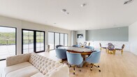 This contemporary single-floor space is thoughtfully designed for entertainment and social gatherings, combining modern aesthetics with versatile functionality. Light hardwood flooring runs throughout, complementing the soft sage-green and crisp white walls that give the interior a clean, sophisticated feel.

The open layout encompasses several distinct zones. A central gaming area anchors the space with a dedicated oval poker table surrounded by plush powder-blue velvet chairs with gold-accented bases, offering a refined yet playful atmosphere. Adjacent to it, a secondary lounge area features a round marble-top table paired with elegant gold-legged velvet chairs in deep purple, accompanied by a built-in white cabinetry unit ideal for storage and serving.

A media lounge area provides comfortable seating with tufted cream sofas arranged around a large wall-mounted flat-screen TV, creating an inviting setting for relaxed viewing. Curated wall art adds character and personality throughout the space.

One of the most striking features is the expansive wall of floor-to-ceiling black-framed glass doors and sliding panels that flood the interior with natural light and open directly onto a spacious outdoor terrace. The terrace overlooks lush, tree-lined scenery, seamlessly blending indoor and outdoor living. The overall ambiance is upscale and modern, designed to accommodate both intimate gatherings and larger social events.