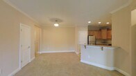 This single-floor space offers a thoughtfully designed open-concept layout that seamlessly connects the living area and kitchen. The spacious living area is finished with soft, plush wall-to-wall carpeting in a warm neutral tone, creating a comfortable and inviting atmosphere. Crown molding runs along the ceiling perimeter, adding a refined architectural detail throughout the space. Recessed lighting, complemented by a central pendant light fixture, provides well-distributed illumination across the room.

The kitchen is positioned adjacent to the living area and features warm maple cabinetry, dark granite countertops with subtle veining, and stainless steel appliances, including a full-size refrigerator. A peninsula with an extended countertop overhang serves as a natural divider between the kitchen and living space, offering casual seating and additional prep surface area.

The neutral, cream-toned walls throughout lend a clean and versatile canvas, allowing for a wide range of interior styling. Multiple doorways are visible, indicating access to additional rooms and storage. The overall ambiance of the space is bright, airy, and move-in ready, with classic finishes that balance practicality and understated elegance.