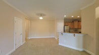This single-floor space offers a thoughtfully designed open-concept layout that seamlessly connects the living area and kitchen. The spacious living area is finished with soft, plush wall-to-wall carpeting in a warm neutral tone, creating a comfortable and inviting atmosphere. Crown molding runs along the ceiling perimeter, adding a refined architectural detail throughout the space. Recessed lighting, complemented by a central pendant light fixture, provides well-distributed illumination across the room.

The kitchen is positioned adjacent to the living area and features warm maple cabinetry, dark granite countertops with subtle veining, and stainless steel appliances, including a full-size refrigerator. A peninsula with an extended countertop overhang serves as a natural divider between the kitchen and living space, offering casual seating and additional prep surface area.

The neutral, cream-toned walls throughout lend a clean and versatile canvas, allowing for a wide range of interior styling. Multiple doorways are visible, indicating access to additional rooms and storage. The overall ambiance of the space is bright, airy, and move-in ready, with classic finishes that balance practicality and understated elegance.
