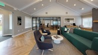 This thoughtfully designed single-floor space combines contemporary aesthetics with functional versatility. The main lounge area immediately draws attention with its dramatic vaulted ceilings accented by exposed white-painted beams and a striking sputnik-style chandelier. A floor-to-ceiling tile-clad fireplace serves as a focal point, flanked by a wall-mounted television and a warm wood hearth shelf. The seating arrangement features a bold emerald green sectional sofa paired with charcoal sculptural accent chairs and a round marble-top coffee table, all anchored by a neutral area rug.

Herringbone-patterned hardwood flooring flows throughout, lending a sophisticated visual texture to the space. An abundance of natural light pours in through multiple large windows fitted with white horizontal blinds, and French doors provide a seamless connection to the outdoors.

A sleek glass-and-black-steel partition wall defines a dedicated workspace area, furnished with desks and task chairs, offering privacy while preserving an open, connected feel. An adjacent corridor leads to additional rooms, maintaining a smooth and intuitive layout. The overall ambiance is modern, airy, and professionally curated.