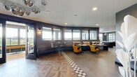 This sophisticated rooftop amenity space combines bold interior design with open-air outdoor access, creating a dynamic environment suited for both casual gatherings and social events. The interior is defined by striking design choices, including a geometric patterned tile floor that transitions between a checkerboard motif and a wood-inlay parquet design, adding visual depth and character throughout. Dark charcoal walls contrast beautifully with bright, curated furnishings — most notably a bold yellow curved sofa, velvet amber accent chairs, and eclectic occasional seating in rich earth tones. A statement pendant light with a sculptural metallic shade anchors the lounge area, while cluster globe disco-ball chandeliers lend a playful, upscale energy to the dining and seating zones. Built-in banquette seating lines the windowed walls, bathing the space in natural light and offering sweeping outdoor views. A sage-green open shelving unit displays decorative objects alongside a wall-mounted TV, while textured wallcoverings and a floral-print ceiling add artistic flair. The adjacent outdoor terrace features warm wood decking, a projector, lush planters, and open-railing views of the surrounding skyline — offering an inviting extension of the interior for al fresco entertaining.