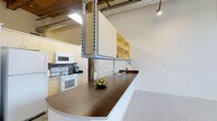 This 1-bedroom, 1-bathroom unit showcases an industrial-modern aesthetic, blending raw architectural elements with functional design. The open-concept layout is immediately striking, highlighted by soaring ceilings with exposed silver ductwork and structural steel supports that lend the space an urban, loft-like character. Track lighting and a skylight contribute to a bright, well-lit environment throughout.

The kitchen is a central feature, anchored by a generously sized curved peninsula with a rich dark wood countertop and an integrated sink. White cabinetry and appliances, including a range and over-the-range microwave, provide a clean contrast against the industrial backdrop. Open shelving in warm wood tones adds both storage and visual warmth to the upper walls.

Speckled light-colored flooring runs throughout, offering durability and a neutral foundation that complements the space's mixed materials. Original wooden structural posts add a historic touch, hinting at the building's character and past. The overall ambiance balances utilitarian industrial design with comfortable, livable details, creating a distinctive and functional living environment.