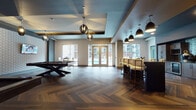 Located in the heart of Charlotte's bustling Trade Street corridor, this upscale amenity lounge delivers a refined, social atmosphere designed for leisure and entertainment. The space is anchored by rich herringbone-patterned hardwood flooring that flows throughout, lending warmth and visual depth to the contemporary interior.

The billiards area serves as a central focal point, featuring multiple pool tables accented by sculptural pendant lighting and textured geometric wall panels. Wall-mounted flat-screen televisions provide additional entertainment throughout. A dedicated wet bar area offers dark cabinetry with glass-front uppers, a striking metallic brick tile backsplash, and stainless steel appliances including a built-in microwave, wine cooler, and oven — ideal for hosting and entertaining.

Bar-height seating with sleek white upholstered chairs surrounds a dark central island, while an intimate lounge nook outfitted with emerald green velvet chairs, hammered gold accent tables, and geometric pendant lighting provides a more relaxed retreat. Glass partitions and sliding doors define this cozy lounge area while maintaining visual openness.

Large floor-to-ceiling windows and glass French doors bathe the space in natural light and offer direct views of an outdoor pool area, seamlessly connecting the interior to the exterior. The layered ceiling design with contrasting teal and warm tones adds architectural interest throughout.