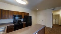 This single-floor space includes a well-appointed kitchen that serves as a central feature of the layout. The kitchen is equipped with a full suite of black appliances, including a refrigerator, electric range, and over-the-range microwave, all complemented by warm, medium-toned wood cabinetry that provides ample upper storage. The countertops feature a speckled neutral finish that adds visual texture while remaining practical for everyday use. A peninsula extends from the main counter, offering additional prep space and the potential for casual seating, subtly separating the kitchen from the adjacent living area. Recessed lighting and a flush-mount ceiling fixture provide layered illumination throughout the space. The flooring transitions between tile and hardwood-style material, adding dimension to the overall design. Two paneled doors are visible in the background, suggesting access to additional rooms or storage within the unit. The neutral wall tones and clean lines throughout contribute to a straightforward, functional ambiance suited to comfortable daily living.