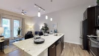 This single-floor residence showcases a thoughtfully designed open-concept layout that integrates the kitchen and living areas into one cohesive, functional space. The kitchen serves as a central highlight, featuring a generously sized island topped with striking granite countertops displaying characteristic white and grey veining. Dark espresso cabinetry provides a bold contrast against the light stone surfaces, while a stainless steel undermount sink adds a clean, contemporary touch. Overhead, a combination of track lighting and elegant pendant lights illuminate the workspace, creating both practical task lighting and an inviting atmosphere.

The light-toned hardwood-style flooring extends throughout, unifying the kitchen and living areas while contributing to the space's bright, airy feel. Smooth, neutral-toned walls enhance the sense of openness, and the ceiling fan visible in the living area suggests comfort-focused design. The adjacent living area is visible beyond the island, reinforcing the open-plan concept. Overall, the space balances modern aesthetics with everyday functionality, offering a welcoming and well-appointed interior environment.