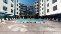 Located at 1018 N Caldwell St in Charlotte's NoDa neighborhood, this outdoor amenity level showcases a thoughtfully designed courtyard retreat enclosed by a striking multi-story residential building with a bold navy and white exterior. At the heart of the space is a generously sized swimming pool with crystal-clear water, flanked by a broad concrete sun deck furnished with lounge chairs, umbrellas, and in-pool seating platforms perfect for relaxing in the Carolina sun. Tropical palm trees are strategically placed throughout the courtyard, adding a resort-like ambiance to the urban setting. A modern steel pergola structure runs along the perimeter, providing shaded transitional zones that connect the pool deck to the interior amenity spaces. These covered outdoor areas are equipped with contemporary modular seating arrangements and a ping pong table, offering versatile spaces for both casual socializing and active recreation. Floor-to-ceiling glass sliding doors allow seamless indoor-outdoor flow between the pergola-covered terrace and the interior lounge visible beyond. The polished large-format concrete tile flooring, sleek glass pool barriers, and refined black metal framework throughout lend the entire amenity deck a cohesive, upscale aesthetic well-suited to modern urban living.