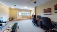Located in Jacksonville, FL's 32256 business district, this single-story business center offers a well-organized and functional workspace designed to support professional operations. The interior features neutral beige walls, textured ceilings, and wall-to-wall carpeting throughout, creating a cohesive and professional atmosphere.

The main work area is equipped with multiple computer workstations arranged along wood-toned desks with ergonomic office chairs, providing ample space for productivity. A ceiling fan with a light fixture overhead ensures comfortable air circulation, while a multi-pane window near the entry door allows natural light to fill the workspace. Additional ambient lighting is provided through recessed fixtures and a table lamp positioned near a wooden storage cabinet.

A dedicated reception and waiting area features sleek black leather seating, including a sofa and accent chairs, arranged around a wooden coffee table. Crown molding accents the ceiling in this area, adding a polished touch, and a large landscape-inspired wall canvas enhances the welcoming ambiance. The space also includes restroom access directly off the waiting area, contributing to a complete, self-contained professional environment suited for a variety of business functions.