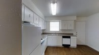 This single-story space includes a well-organized kitchen that serves as a central feature. The kitchen is designed with ample white cabinetry lining both the upper and lower walls, providing generous storage throughout. Countertops in a neutral tone offer practical workspace, complemented by a double-basin sink positioned beneath a window that allows natural light to filter in. The appliance suite includes a refrigerator, dishwasher, and range, all finished in white and black tones that coordinate with the clean, bright aesthetic of the cabinetry. The flooring throughout features warm wood-style planking, adding a grounded, cohesive look to the space. Overhead, a flush-mount fluorescent light fixture ensures the kitchen remains well-lit. The layout follows an efficient L-shape, maximizing usable space while maintaining clear pathways. The textured ceiling and neutral wall tones contribute to a simple, functional ambiance. Overall, the space presents a straightforward, practical design suited for everyday use.